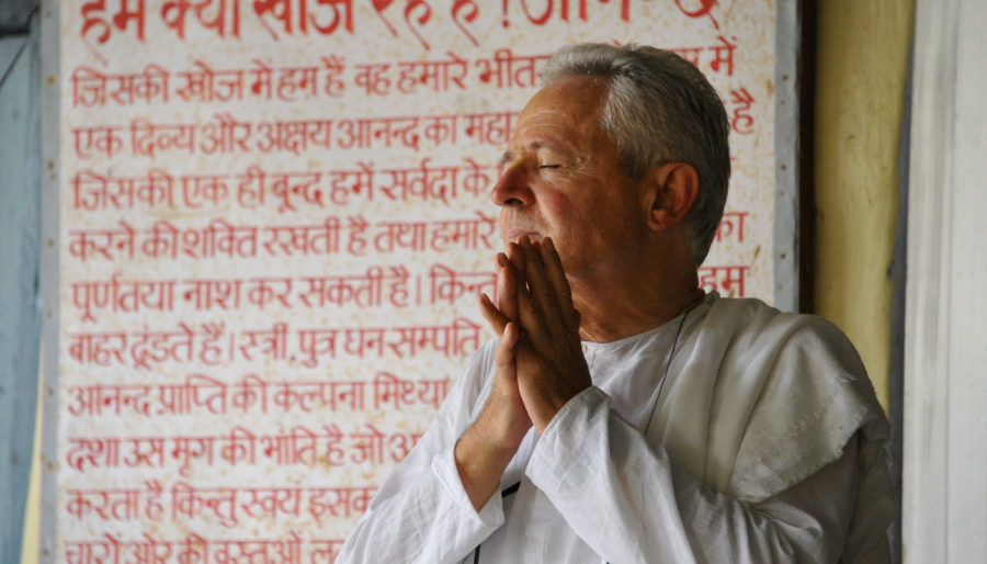 Atma-vidya: la conoscenza del sé. MARCO FERRINI CON MANI GIUNTE E OCCHI CHIUSI sullo sfondo un muro con scritte in sanscrito